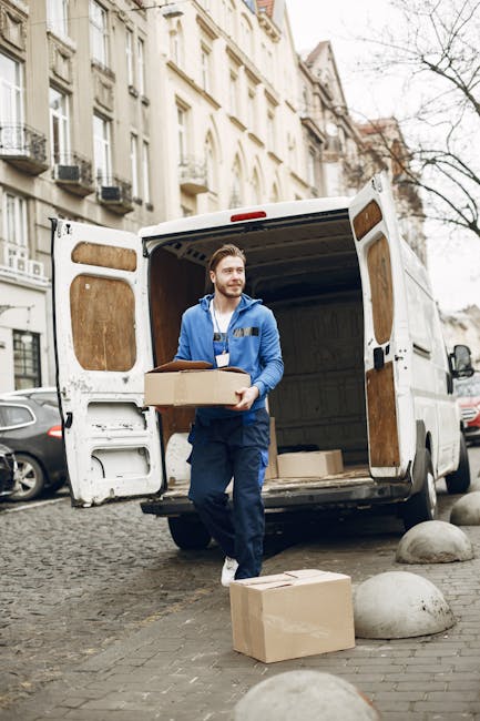 A man wearing a blue jacket and navy work overalls is standing outside on a cobblestone street near a white delivery van, holding a cardboard box in his hands as part of a home relocation or furniture transport process. The van, parked on the street, has its rear doors open, revealing an interior lined with wood panels. Several cardboard boxes are visible around him on the ground, including one resting on a large, rounded concrete bollard. The setting appears to be a residential area with multi-story buildings featuring classic architectural details and balconies. The man is in the midst of loading or unloading boxes, with some on the pavement and others being carried, supporting a professional removals or moving service offered by Man with Van Brompton, as part of a furniture moving or packing and moving operation in Fulham Road.