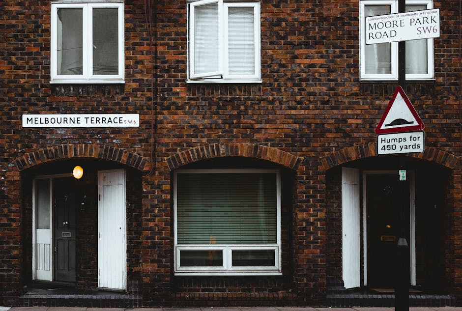 Photograph of a brick residential building on Melbourne Terrace, SW6, with two upper-floor windows fitted with white frames and internal blinds. The building has two entrance doors on the ground floor, each set within an arched brick surround; one door is open and the other closed, both painted in dark colors. A small exterior light is illuminated beside the left door. Above, there are two street signs indicating the location at Moore Park Road and Melbourne Terrace, along with a traffic warning sign about road bumps for 450 yards. The scene takes place during daytime with natural lighting. This setting supports the context of house removals and furniture transport, as part of a home relocation process, with visual details of the building's façade relevant for moving logistics by companies like Man with Van Brompton.