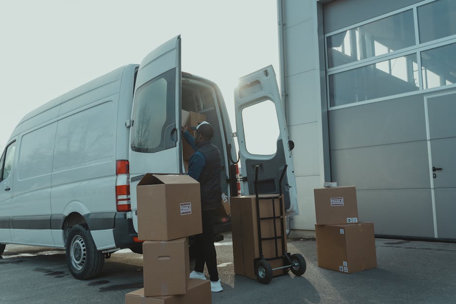 A man wearing a blue jacket and navy work overalls is standing outside on a cobblestone street near a white delivery van, holding a cardboard box in his hands as part of a home relocation or furniture transport process. The van, parked on the street, has its rear doors open, revealing an interior lined with wood panels. Several cardboard boxes are visible around him on the ground, including one resting on a large, rounded concrete bollard. The setting appears to be a residential area with multi-story buildings featuring classic architectural details and balconies. The man is in the midst of loading or unloading boxes, with some on the pavement and others being carried, supporting a professional removals or moving service offered by Man with Van Brompton, as part of a furniture moving or packing and moving operation in Fulham Road.