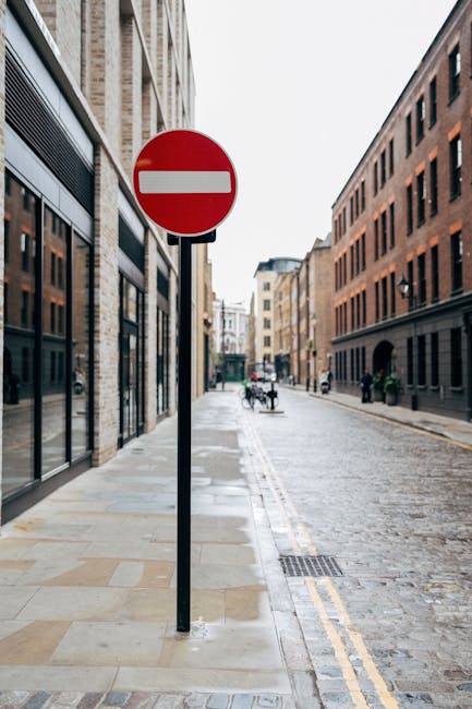 A red circular 'no entry' traffic sign mounted on a black pole is positioned on a cobbled urban street in Old Brompton Road, surrounded by multi-storey brick and stone buildings with large windows. The pavement features a concrete sidewalk with some visible drains, while the narrow road includes double yellow lines along its edge. Pedestrians and cyclists can be seen in the background. This setting is indicative of a typical residential or commercial area suitable for household removals and furniture transport, supporting home relocation services such as those offered by Man with Van Brompton. The environment appears overcast with diffused natural light, emphasizing the functional surroundings for moving logistics and packing activities common during house removals on narrow city streets.