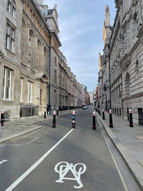 A narrow city street in Old Brompton Road with tall, historic stone buildings on either side, featuring ornate architectural details and multiple chimneys. The street is partially blocked by black and white safety bollards and plastic barriers, preventing vehicle access, while a dedicated bicycle lane with a white painted bicycle symbol runs down the middle. The pavement is clean, and there are some small metal benches and a wheelchair accessible ramp on the left sidewalk. The sky above is cloudy with patches of blue, and the scene appears to be set during daylight hours. This setting illustrates the challenges of home relocation and furniture transport in areas with narrow access, often requiring professional moving services like those provided by Man with Van Brompton, especially when navigating restricted city streets during the loading process of house removals and packing logistics.