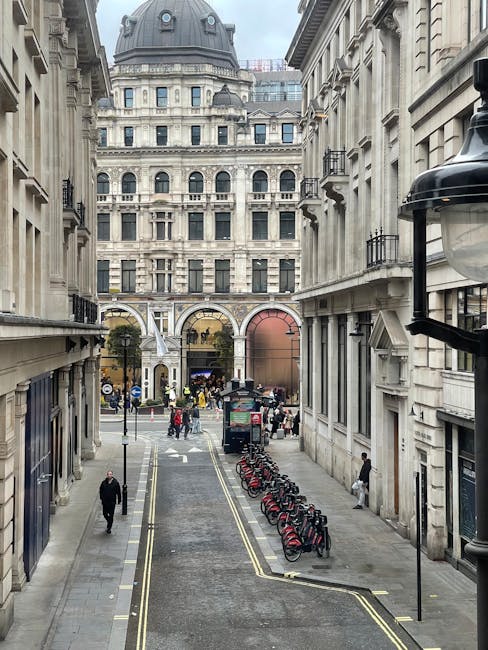 A red circular 'no entry' traffic sign mounted on a black pole is positioned on a cobbled urban street in Old Brompton Road, surrounded by multi-storey brick and stone buildings with large windows. The pavement features a concrete sidewalk with some visible drains, while the narrow road includes double yellow lines along its edge. Pedestrians and cyclists can be seen in the background. This setting is indicative of a typical residential or commercial area suitable for household removals and furniture transport, supporting home relocation services such as those offered by Man with Van Brompton. The environment appears overcast with diffused natural light, emphasizing the functional surroundings for moving logistics and packing activities common during house removals on narrow city streets.