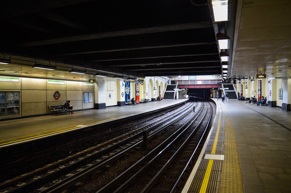 A man in dark clothing, including a black coat and hat, is walking along the pavement outside a modern building near South Kensington underground station in London. The building features large, vertic